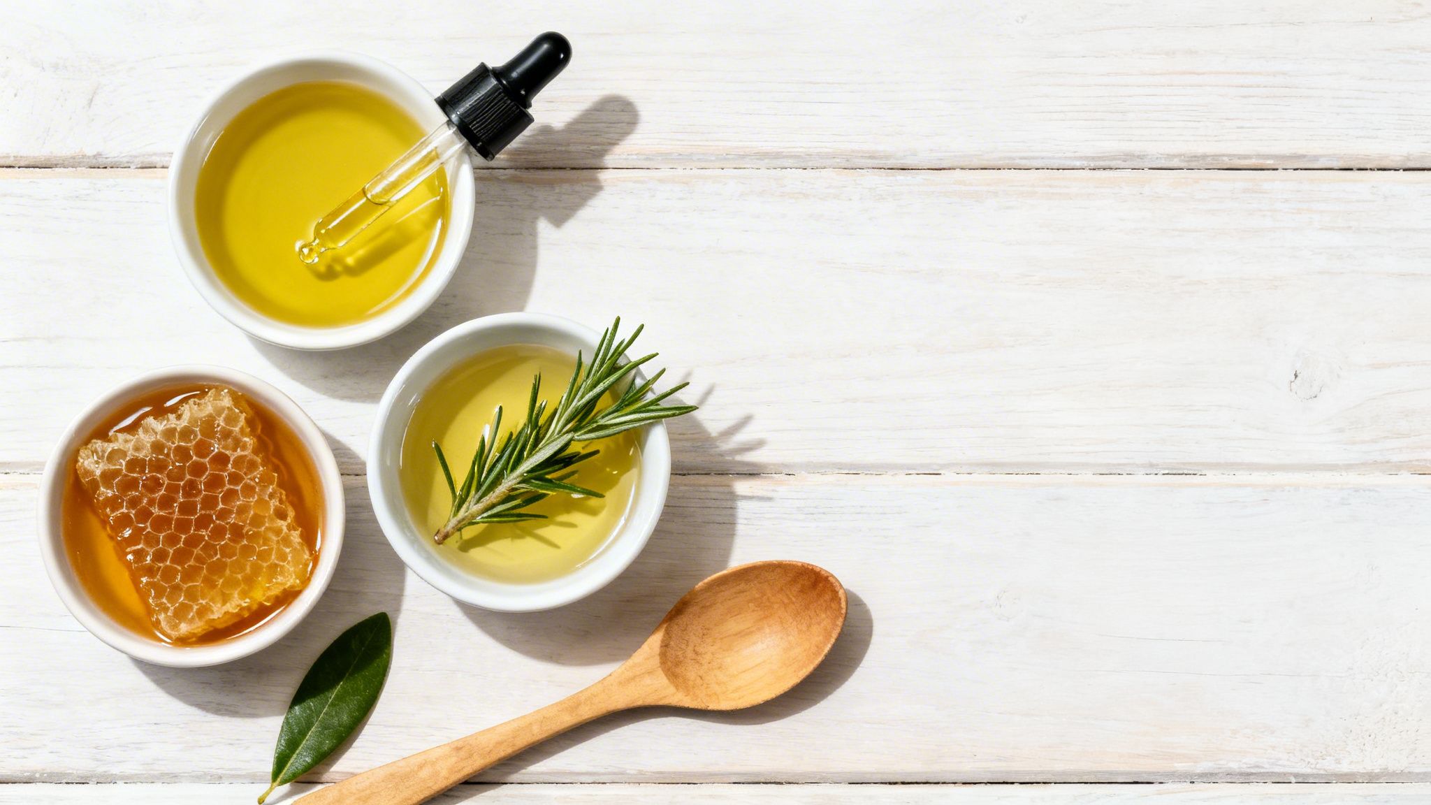 Overhead view of bowls with natural ingredients: olive oil with dropper, honey with honeycomb, and rosemary oil.